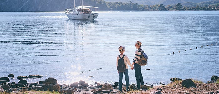 Two people stand on a lake shore, with a boat in the background, highlighting international tourism